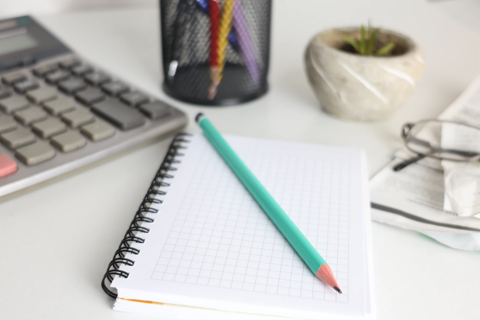 A calculator, open notebook, pencil and small plant arranged on a desk.