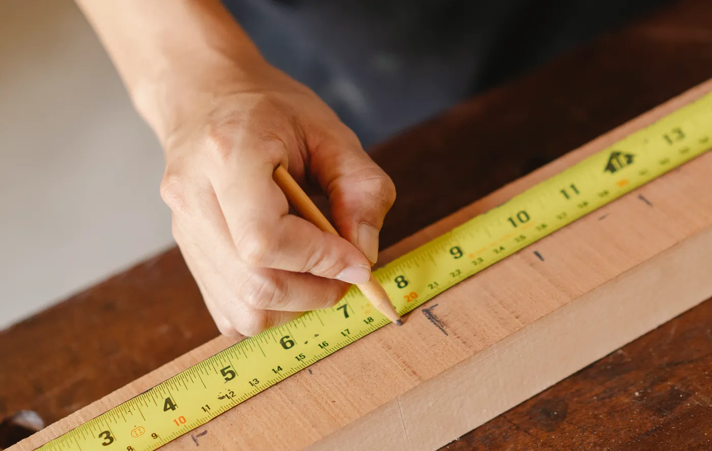 A hand marking a timber plank with a pencil beside a measuring tape.