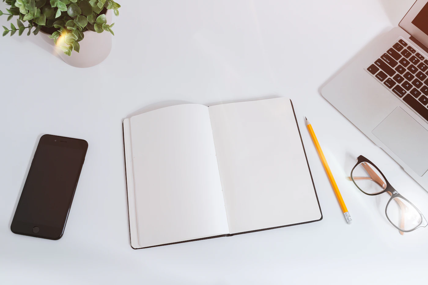 An empty notebook and pencil beside a laptop and phone on a desk.